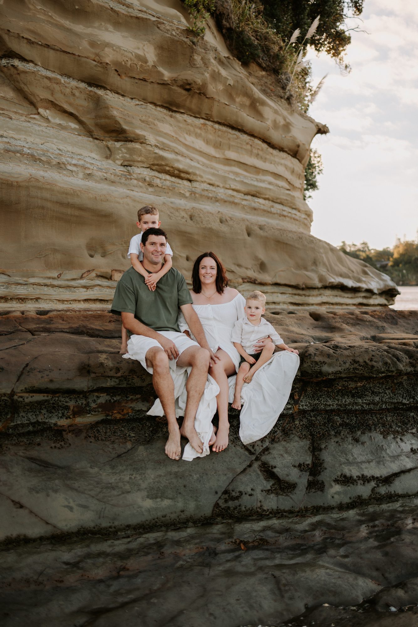 Family of four sitting together on a sandstone cliff ledge at the beach, smiling at the camera