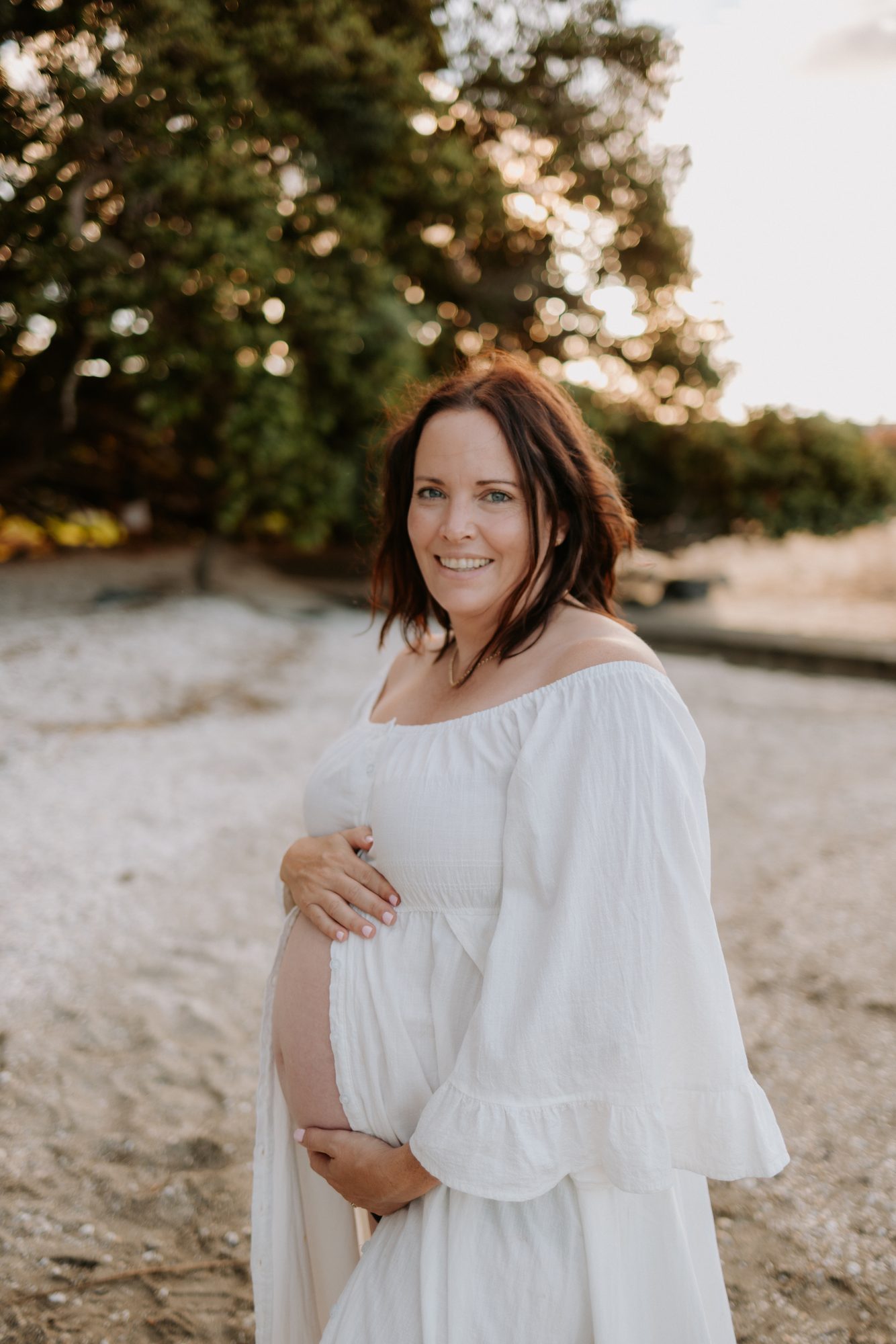Pregnant woman in a white dress cradling her belly under trees at golden hour