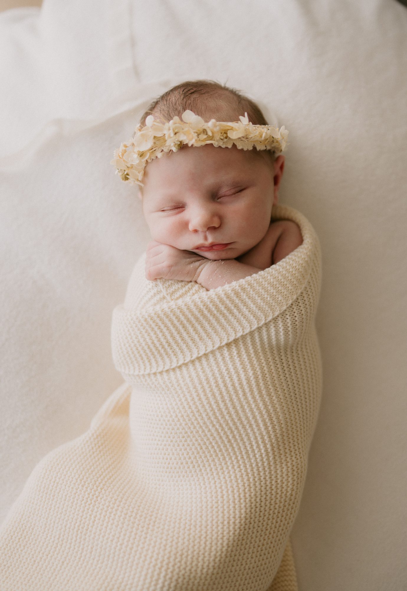 Sleeping newborn baby swaddled in a cream wrap, wearing a dried flower crown