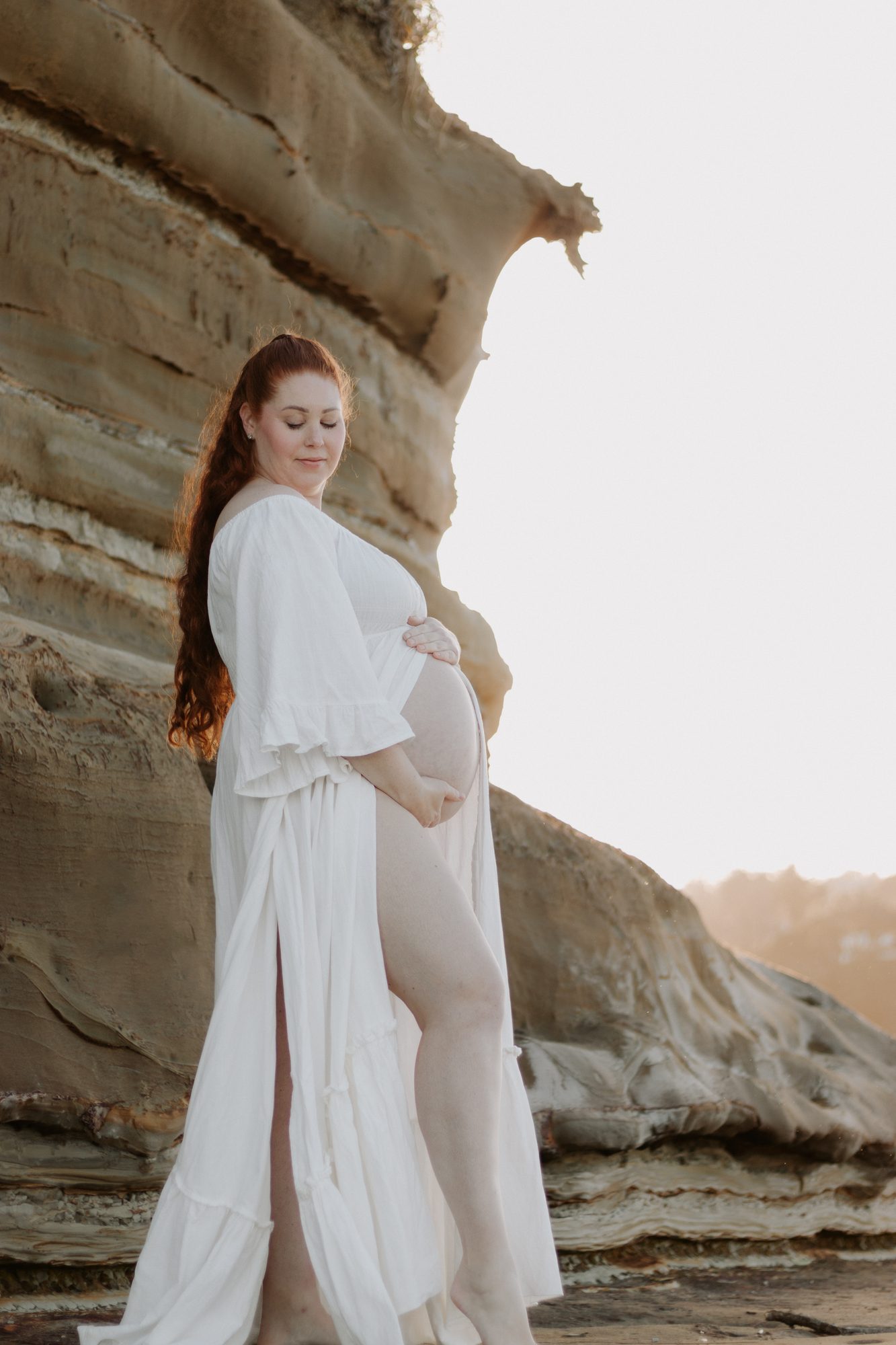 Pregnant woman in a white dress standing side-on against a sandstone cliff